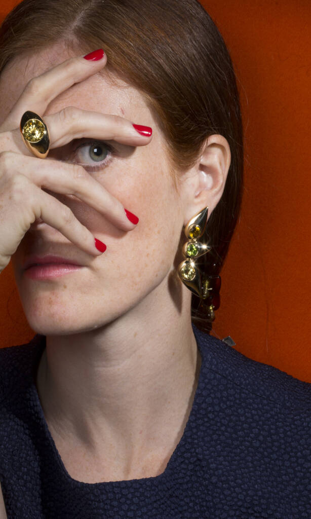 woman with hand over face with red nails wearing a ring and earrings against an orange background